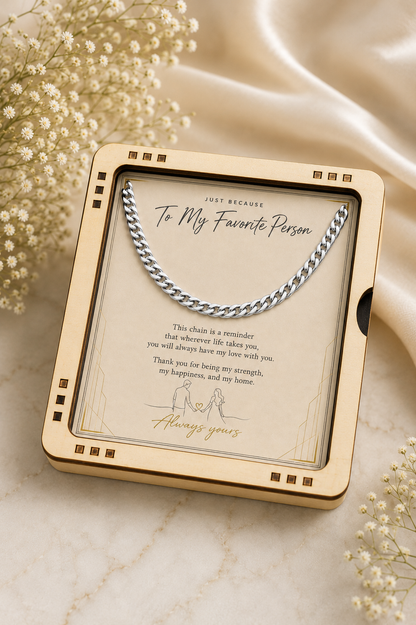 A silver chain necklace displayed inside a square cream-colored gift box with a clear front cover and printed sentimental message, styled on a light marble surface with soft satin fabric and small white flowers around it.