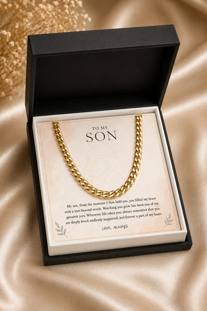 A gold chain necklace presented in a black gift box with a cream insert card addressed 'TO MY SON' and a sentimental message, photographed on a beige satin fabric background.