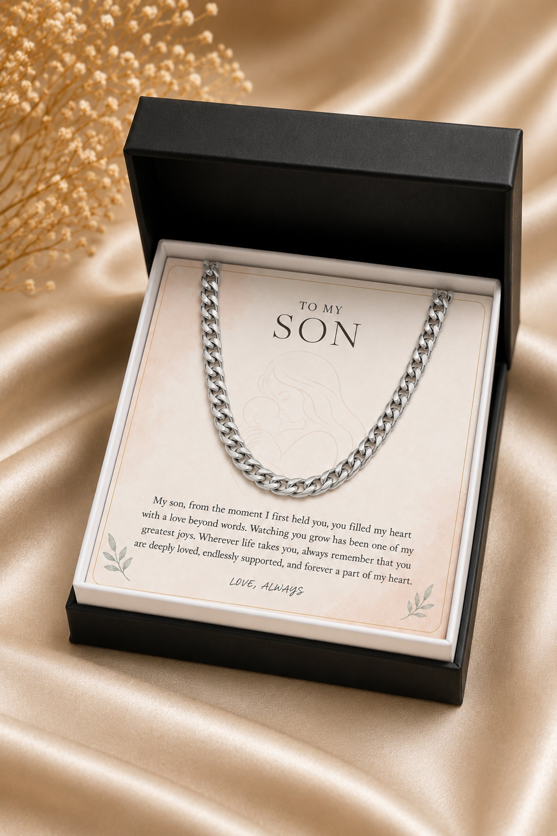 A silver chain necklace presented in an open black gift box with a printed message card addressed 'TO MY SON,' placed on a draped champagne satin fabric background.