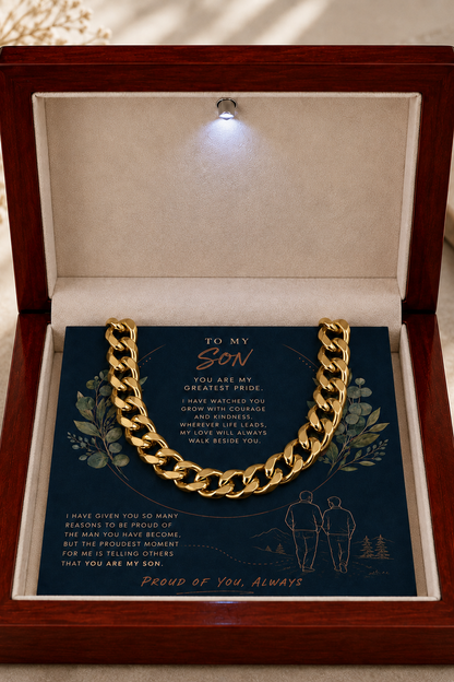 A thick gold Cuban link chain necklace is presented inside an open wooden gift box with a soft cream lining. The chain rests on a dark blue printed card with sentimental text and illustrations, and a small built-in light shines into the box from above.