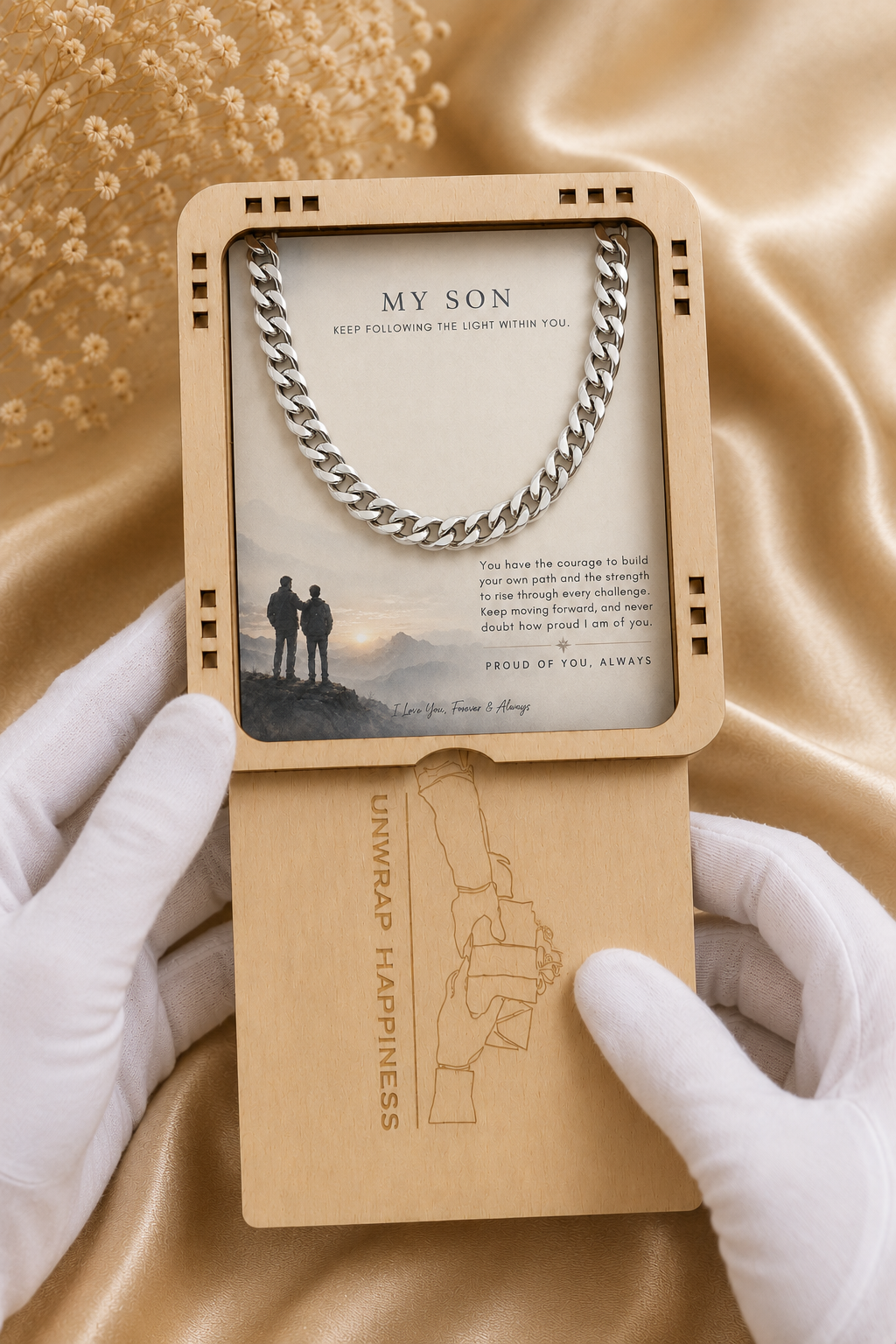 A silver chain necklace presented in a wooden gift box with a printed card that reads 'MY SON' and a message, held open by white-gloved hands over a gold satin fabric background.