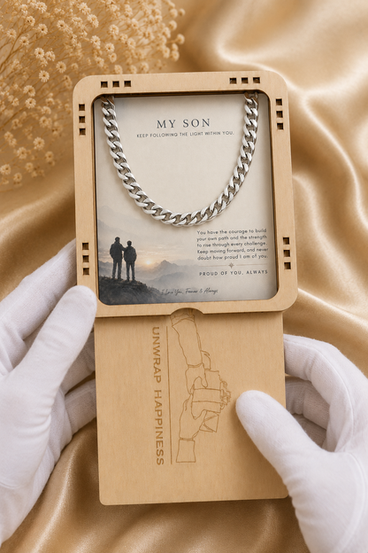 A silver chain necklace presented in a wooden gift box with a printed card that reads 'MY SON' and a message, held open by white-gloved hands over a gold satin fabric background.