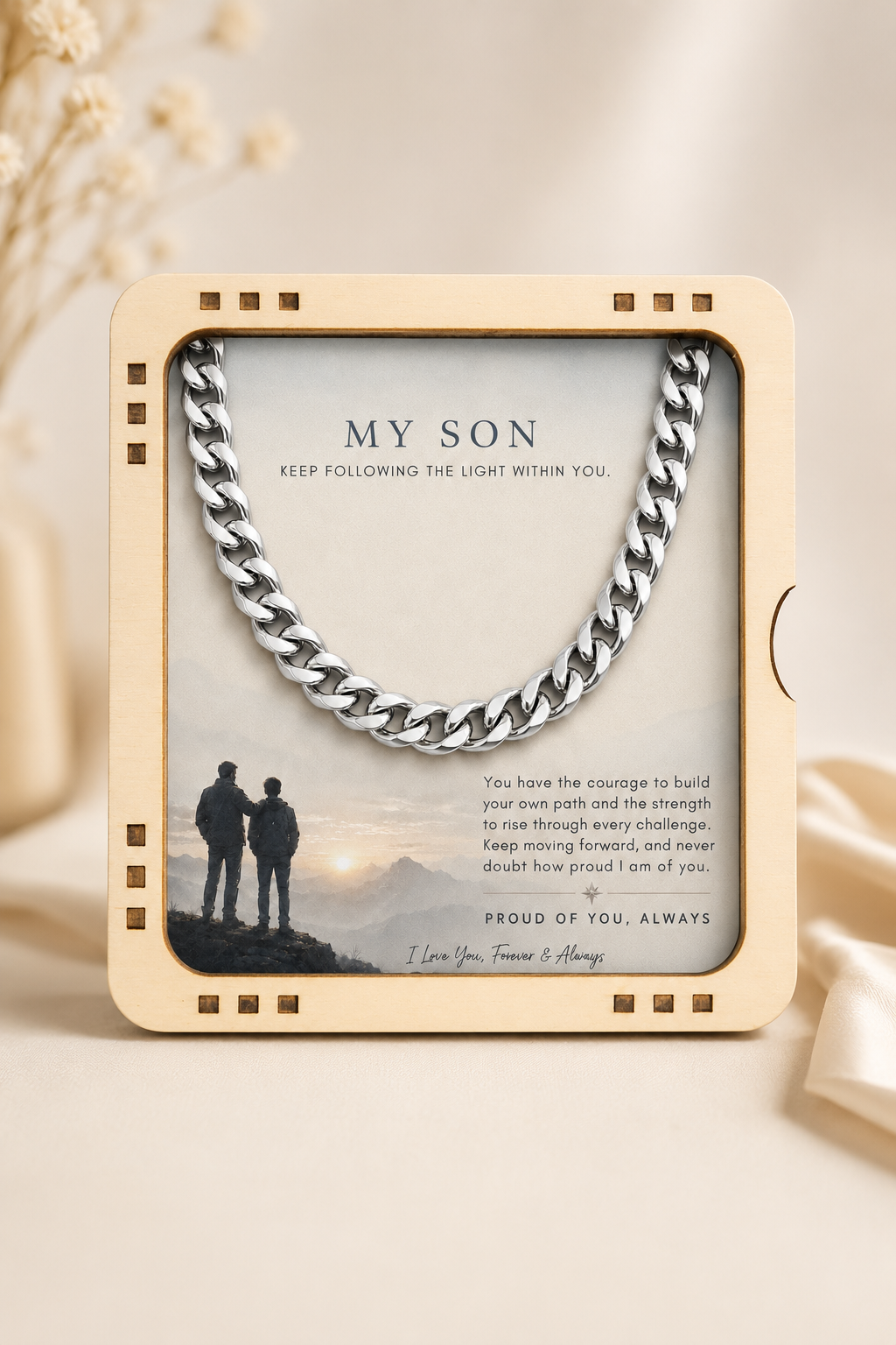 A silver curb chain necklace presented inside a square cream-colored wooden keepsake frame with a sentimental message for 'My Son' printed on the insert card, set against a warm beige background with soft dried flowers.