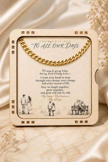 A square cream-colored keepsake photo frame with a gold chain draped across the top, displayed on a beige marble surface with soft cream-colored dried flowers and draped satin fabric in the background. The frame contains a printed sentimental message with illustrations of couples.