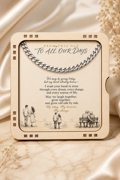 A square cream-colored keepsake box or photo frame with a silver chain across the top, featuring engraved black text reading 'FROM THIS DAY TO ALL OUR DAYS' and sentimental lettering, styled on a beige satin and marble surface with dried flowers.