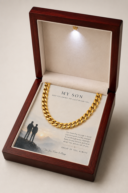 A gold Cuban link chain necklace presented inside an open dark reddish-brown wooden gift box with a soft cream interior. The chain rests on a printed message card addressed 'MY SON' with a small scenic print and sentimental text.