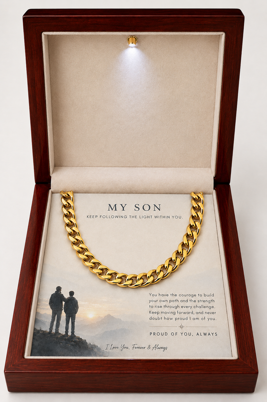 A gold Cuban link chain necklace presented inside an open wooden gift box with a soft cream lining and a printed message card addressed 'MY SON.' The box includes a small built-in light at the top, and the necklace is displayed prominently against a plain light background.