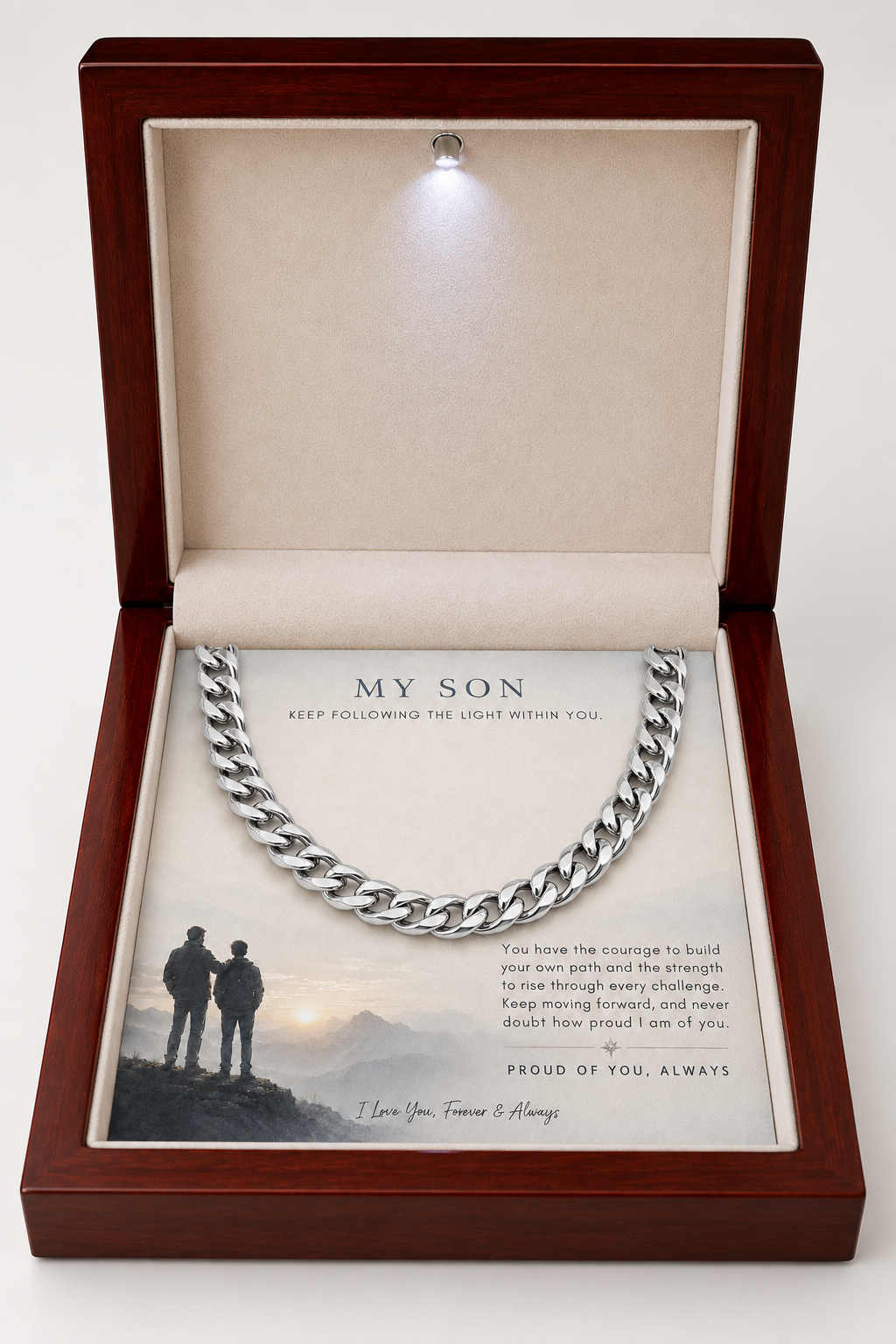 A silver Cuban link chain necklace presented inside an open dark wood gift box with a beige velvet interior. The necklace is displayed over a printed message card addressed 'MY SON' with a small photo of two people standing on a hill at sunset.