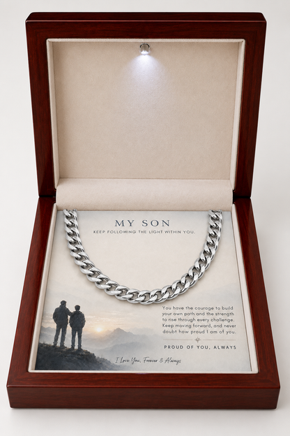 A silver Cuban link chain necklace presented inside an open dark wood gift box with a beige velvet interior. The necklace is displayed over a printed message card addressed 'MY SON' with a small photo of two people standing on a hill at sunset.