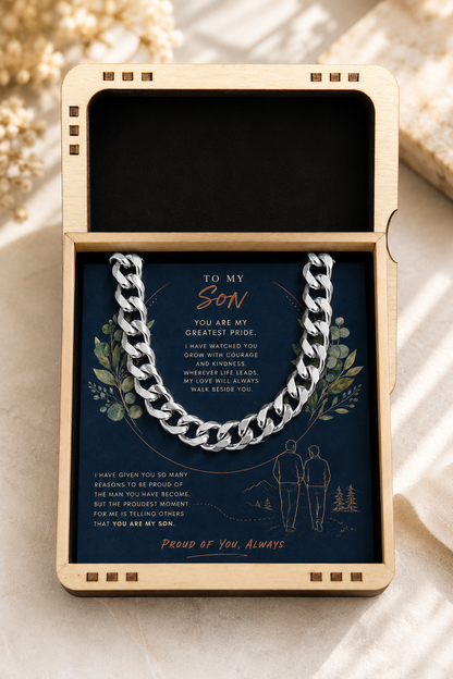 A silver curb chain necklace presented inside an open light wood box with a navy message card addressed to a son, photographed on a soft beige fabric background.
