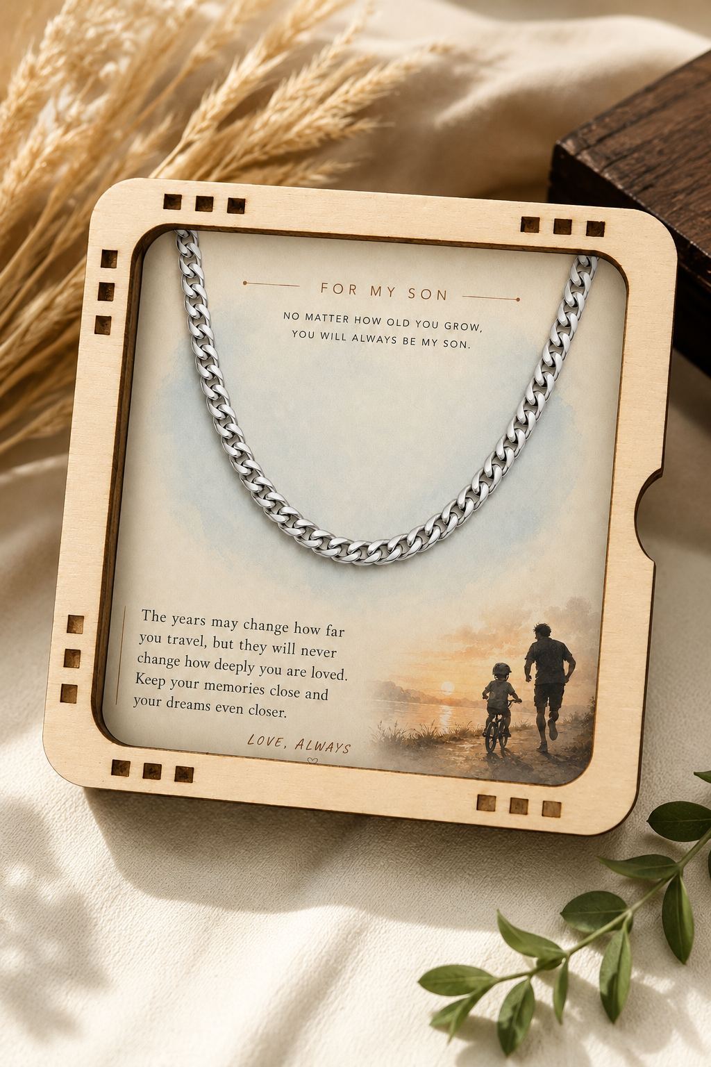 A personalized wooden keepsake frame holding a silver chain necklace and a printed message for a son, styled on a cream fabric with dried wheat and green leaves.