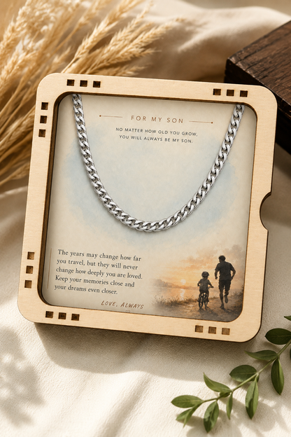 A personalized wooden keepsake frame holding a silver chain necklace and a printed message for a son, styled on a cream fabric with dried wheat and green leaves.