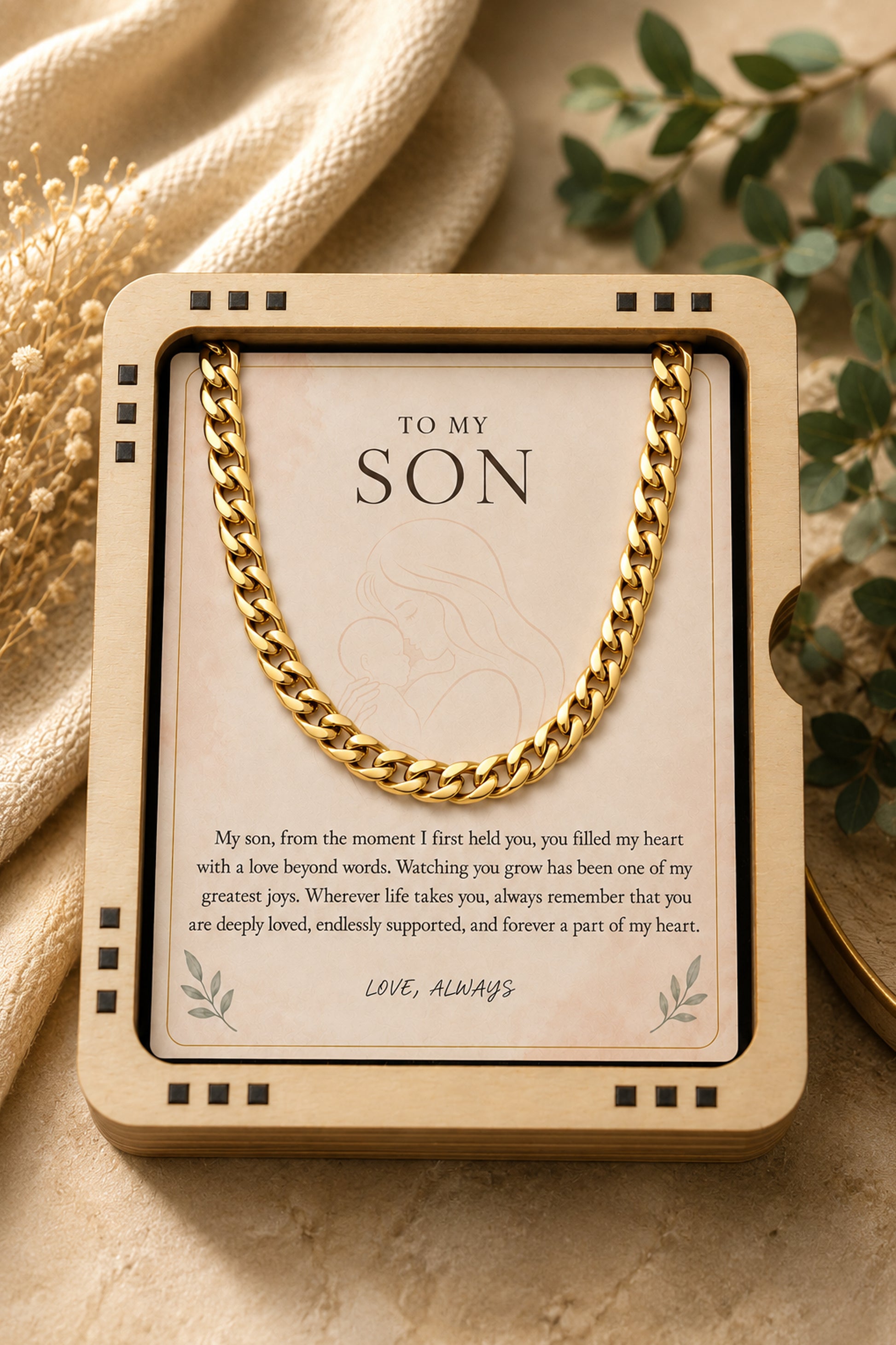 A gold-toned chain necklace displayed inside a square cream gift box with a printed message card addressed 'TO MY SON,' styled on a beige surface with fabric and dried flowers in the background.