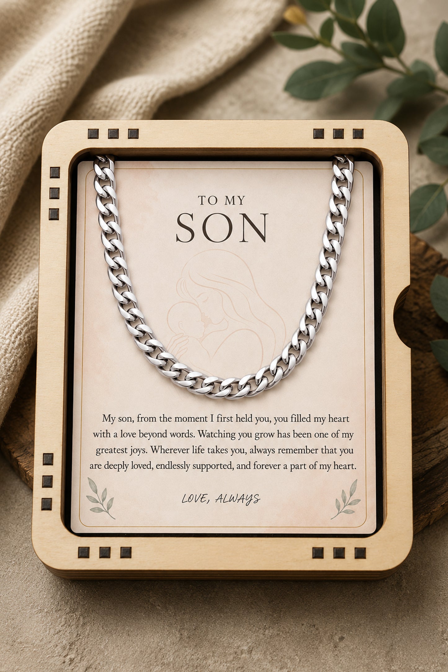 A silver chain necklace presented in a wooden gift box with a printed message card addressed 'TO MY SON,' styled on a neutral fabric surface with soft green leaves in the background.